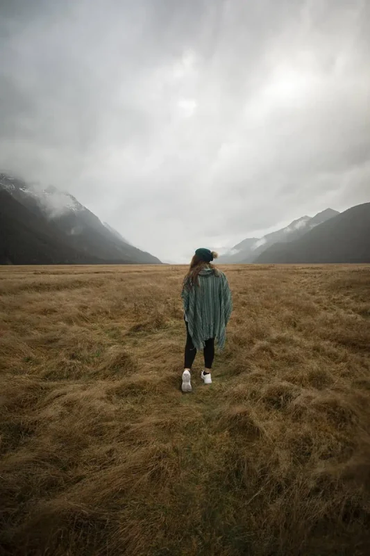 Eglinton Valley: ULTIMATE Guide For Visiting (2025) 5 tasha amy dressed in a green poncho and white sneakers walks across the dry grass, surrounded by towering mountains and an overcast sky. The misty peaks in the distance add to the dramatic scenery of Eglinton Valley.