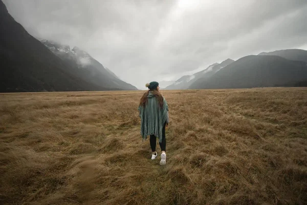 tasha amy dressed in a green poncho and white sneakers walks across a vast, grassy plain under a cloudy sky, with misty mountains in the background. This serene scene is set in Eglinton Valley, New Zealand.