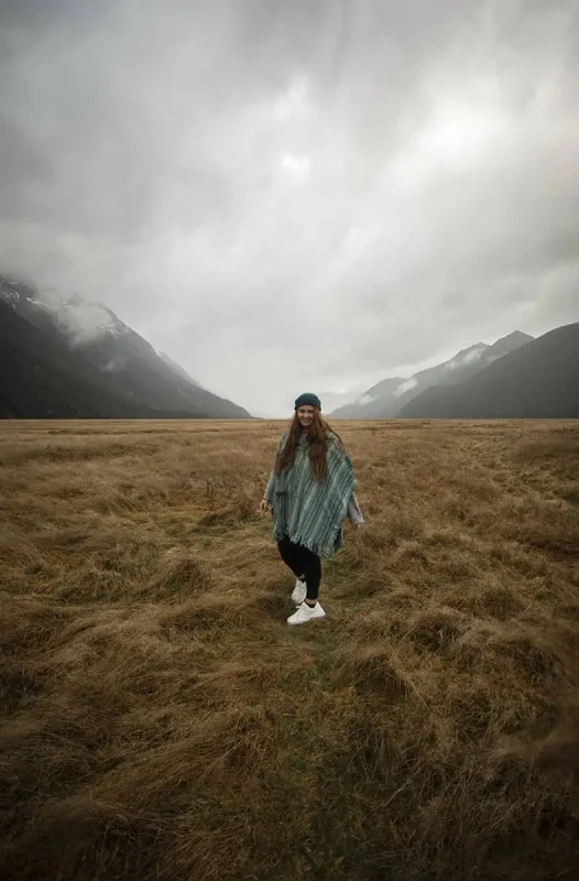 Eglinton Valley: ULTIMATE Guide For Visiting (2025) 4 tasha amy in a green poncho smiles while standing on a vast, grassy plain, with mist-covered mountains in the background. The scene captures the serene and breathtaking landscape of Eglinton Valley.