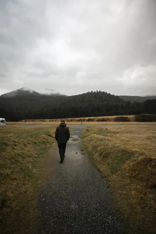 Eglinton Valley: ULTIMATE Guide For Visiting (2025) 12 A person in a dark coat walks along a gravel path leading into the distance, bordered by grassy fields and distant forested hills under a cloudy sky. The peaceful solitude of the scene is accentuated by the misty weather.