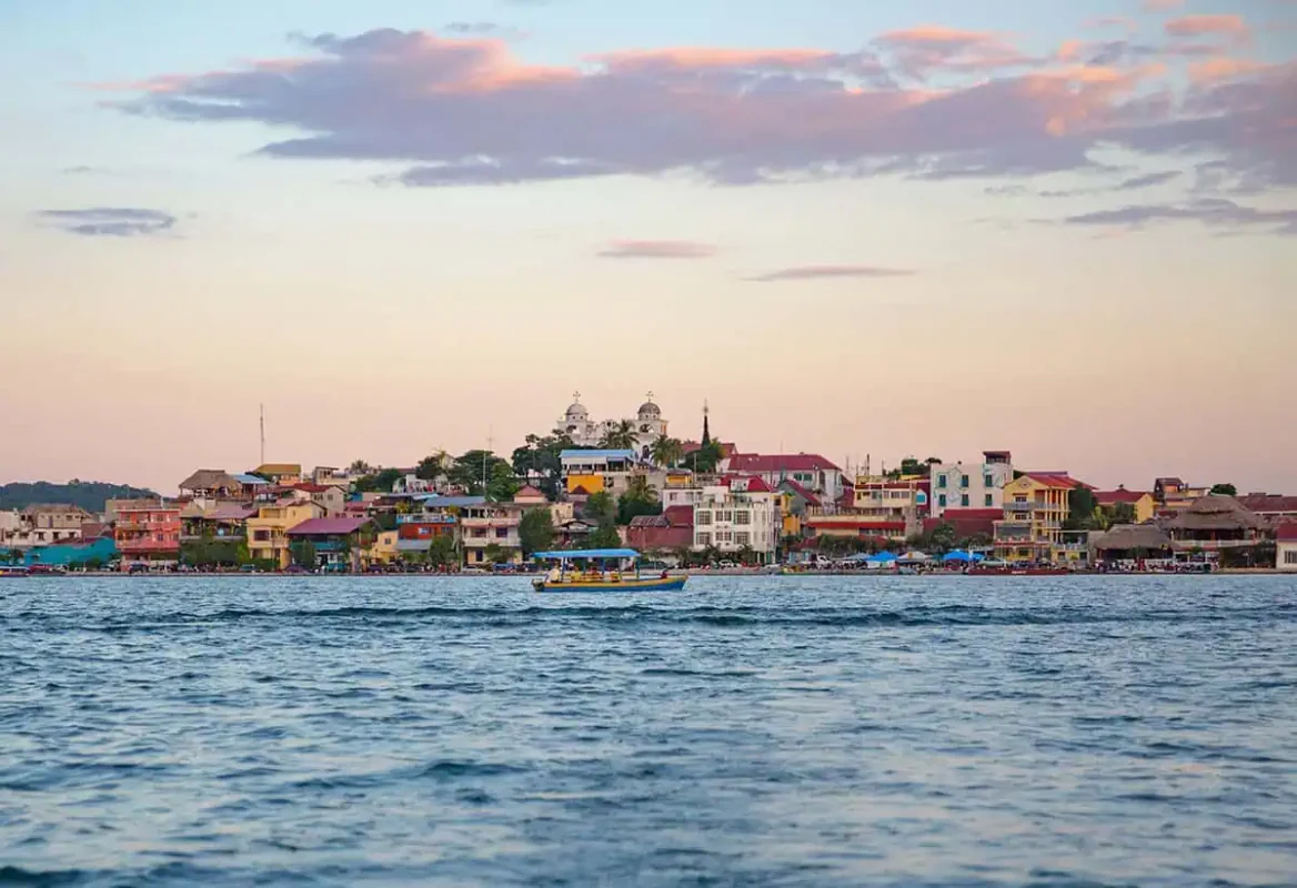 Flores To Belize City: Bus, Shuttle, Or Flight? (2025) 19 The vibrant town of Flores is seen from a distance, with colorful buildings lining the waterfront against a pastel sunset sky. A boat travels across the calm waters, capturing the serene atmosphere of this picturesque Guatemalan town. This image represents the starting point for a journey from Flores to Belize City.