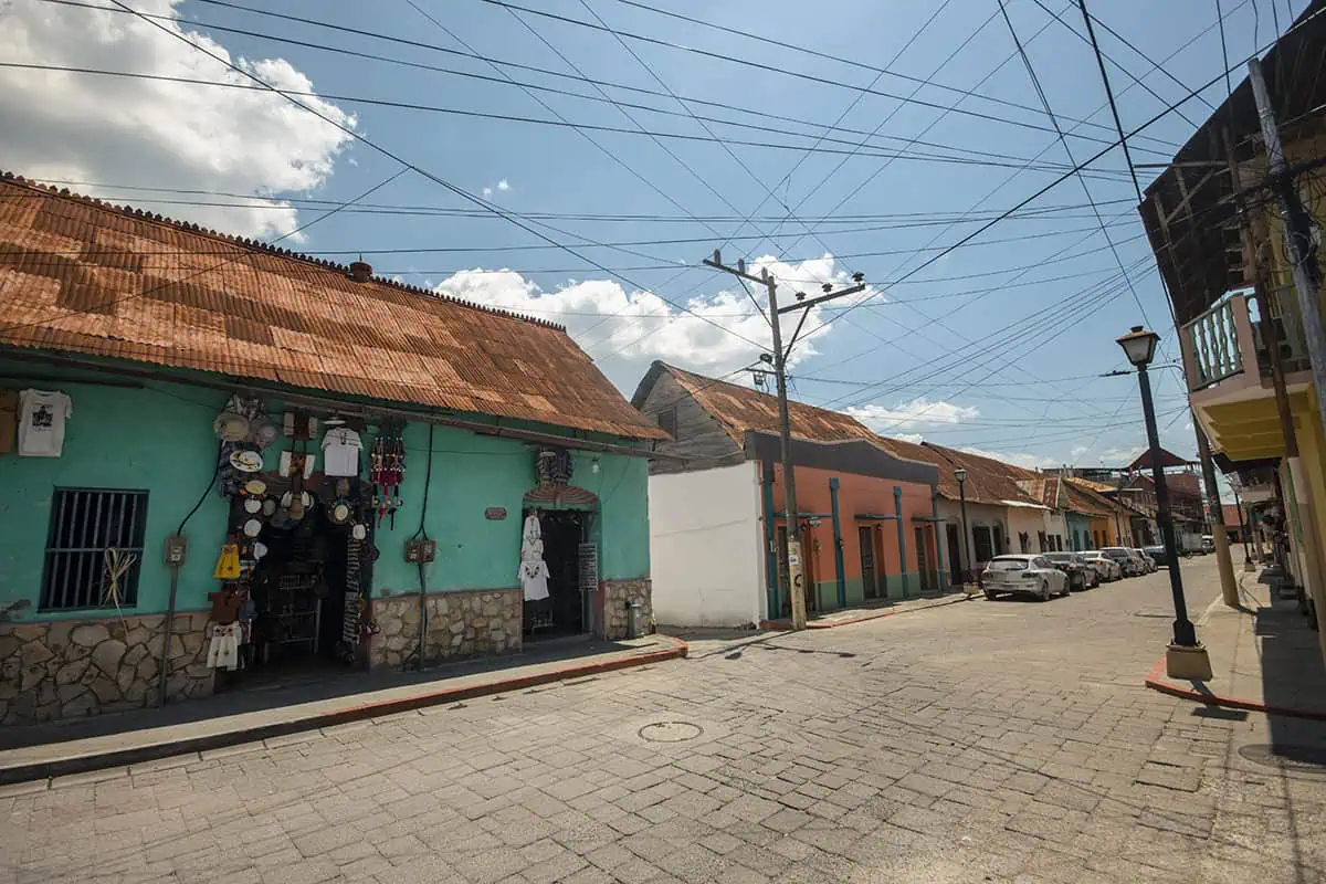 Flores To Belize City: Bus, Shuttle, Or Flight? (2025) 9 Another view of Flores, this time showing a different cobblestone street with a few shops and houses under a clear blue sky. The wires crisscrossing above add a touch of everyday life.