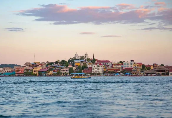 The vibrant town of Flores is seen from a distance, with colorful buildings lining the waterfront against a pastel sunset sky. A boat travels across the calm waters, capturing the serene atmosphere of this picturesque Guatemalan town. This image represents the starting point for a journey from Flores to Belize City.
