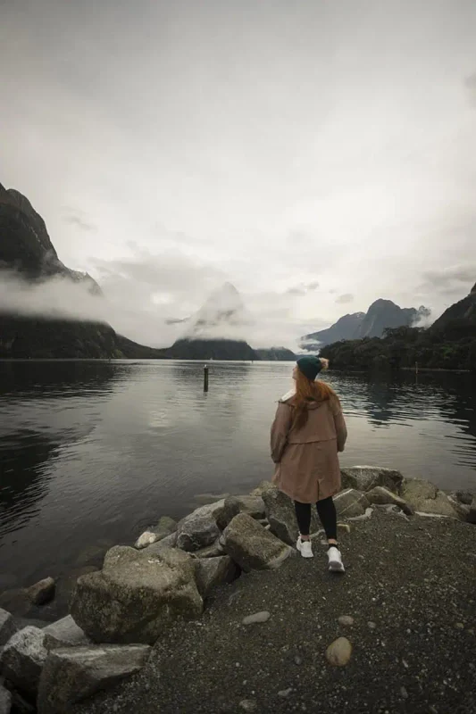 Is Milford Sound Worth It? My Pros & Cons 18 tasha amy standing on the rocky shore with Mitre Peak in the background at Milford Sound.