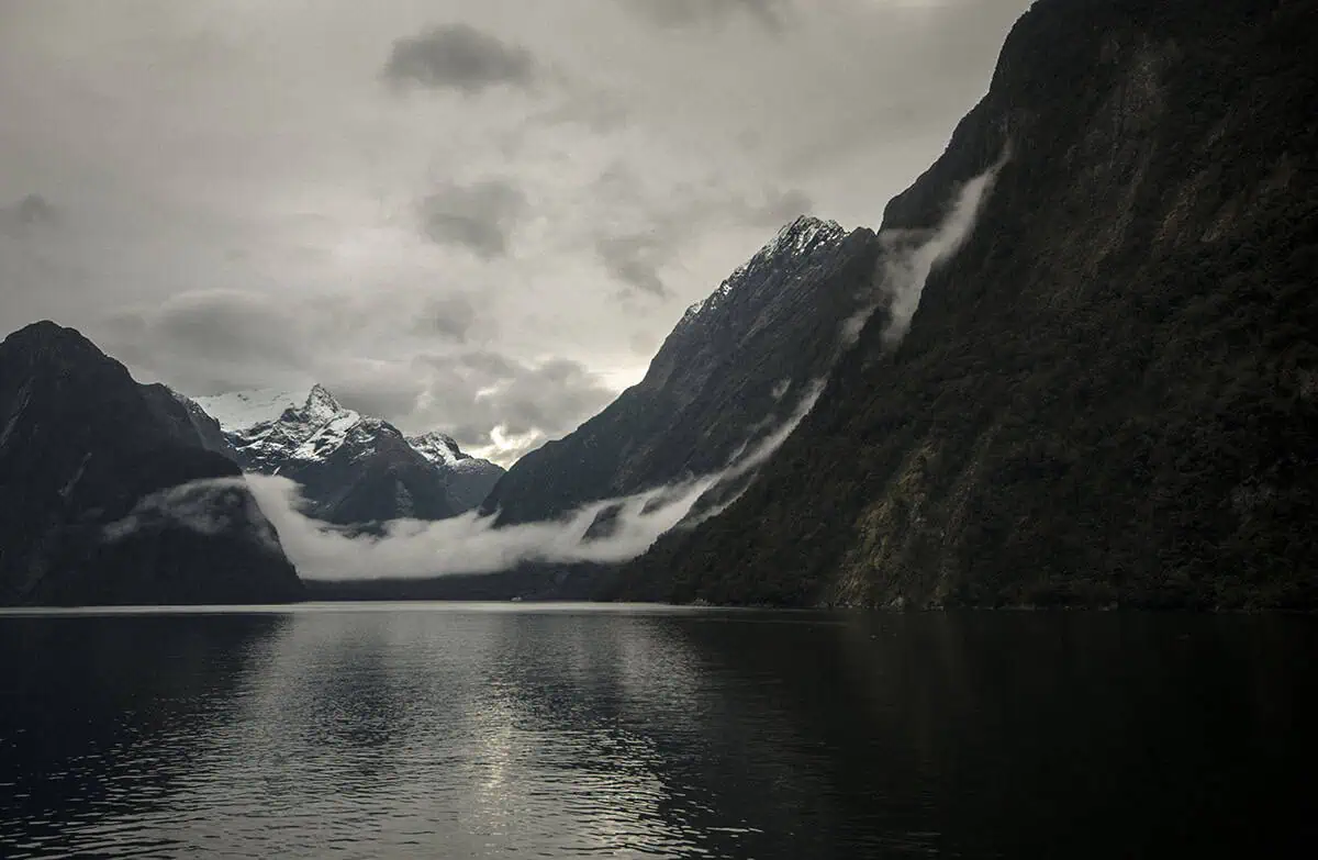Is Milford Sound Worth It? My Pros & Cons 10 Snow covered peaks and low clouds reflected in the calm waters of Milford Sound.