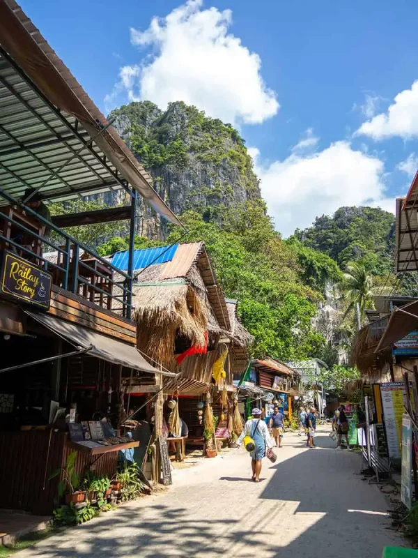 Is Railay Beach Closed: What You Need To Know (2025) 12 Lively street scene at Railay Beach with tourists walking past shops and cafes, set against the backdrop of lush green hills and a clear blue sky.
