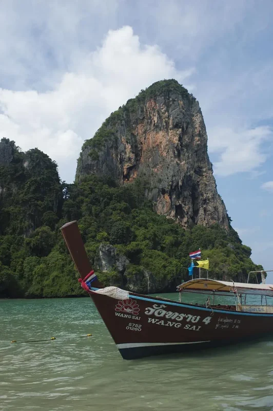 Is Railay Beach Closed: What You Need To Know (2025) 11 A long-tail boat named "Wang Sai" anchored near a towering limestone cliff covered in greenery at Railay Beach. The presence of boats and tourists hints at the beach being operational