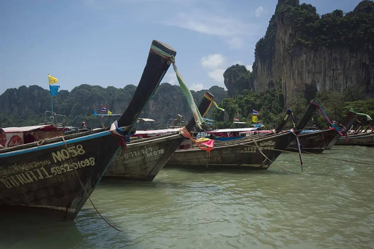 Is Railay Beach Closed: What You Need To Know (2025) 18 Several long-tail boats with colorful flags are lined up in the water with towering cliffs in the background at Railay Beach. This busy scene implies active tourist engagement, questioning if Railay Beach is closed.
