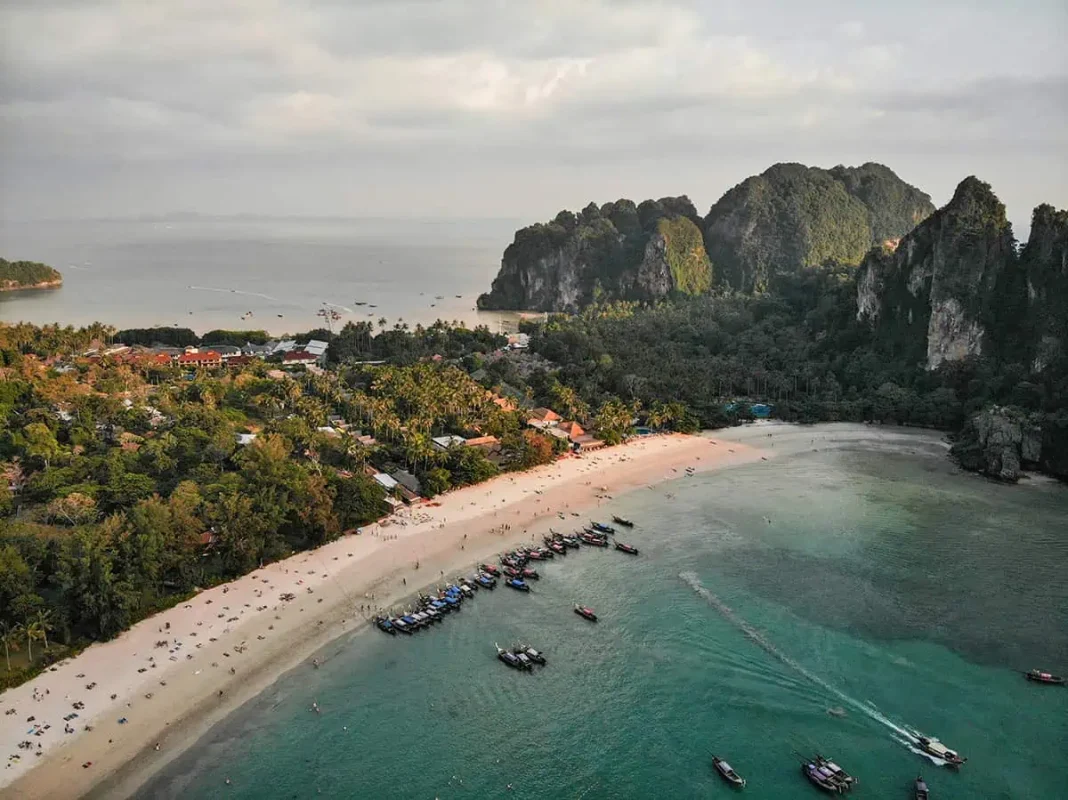 Is Railay Beach Closed: What You Need To Know (2025) 14 Aerial view of Railay Beach with sandy shores, numerous boats docked near the water, and lush greenery surrounding the area under a bright sky.
