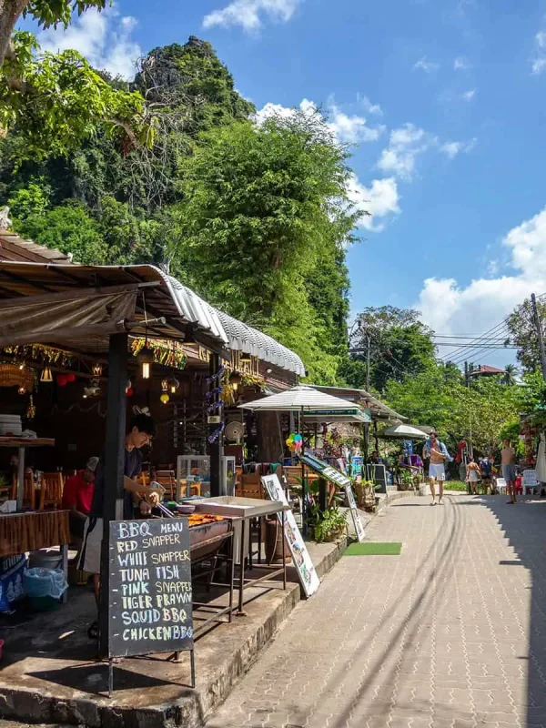 Is Railay Beach Closed: What You Need To Know (2025) 13 A sunny street on Railay Beach lined with small shops and restaurants, featuring a chalkboard menu offering seafood BBQ options like red snapper and tiger prawn.