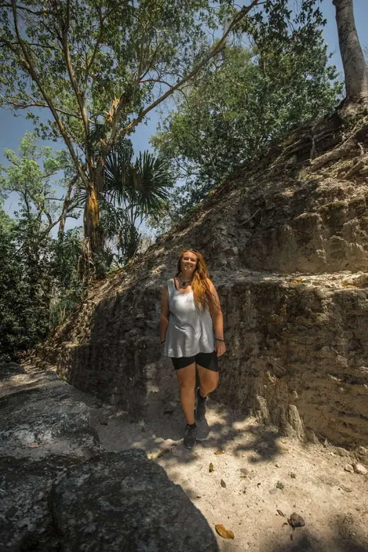 tasha amystanding and smiling on a shaded path beside ancient Mayan ruins surrounded by dense tropical forest. She is wearing a sleeveless top and shorts, capturing the essence of adventure and exploration in belize.