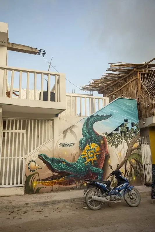 A vibrant mural on a building wall depicting a green alligator with intricate designs on its back. A motorbike is parked in front of the mural, illustrating the urban art scene in Mexico.