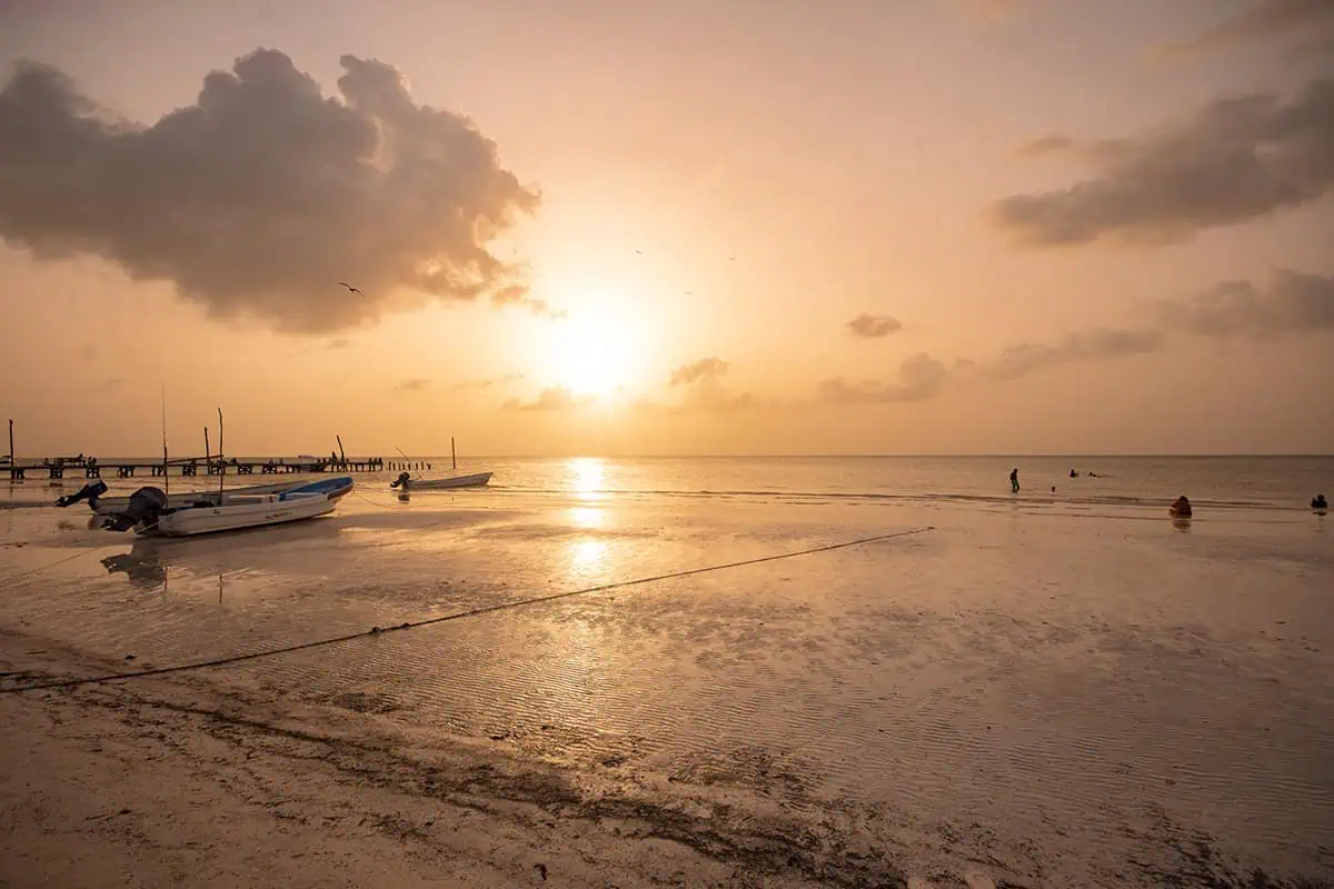 Sunset on an isla holbox beach with small boats anchored in shallow water. The sky is painted with warm hues of orange and pink, reflecting on the water, creating a serene and picturesque moment.