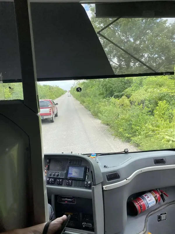 View from inside a bus traveling on a rural road flanked by lush green vegetation. The bus dashboard is visible, and a red car can be seen ahead on the road, highlighting the journey through Mexico and Belize.
