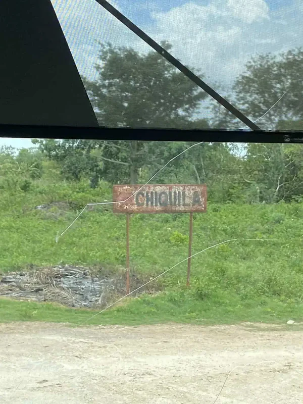 A rusty sign reading "Chiquila" partially obscured by greenery, seen through a bus window, marking a point of interest on a Mexico and Belize itinerary.