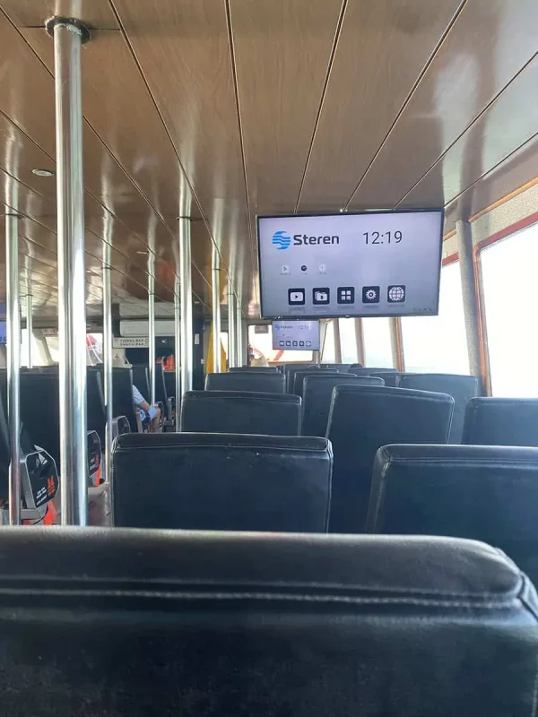 The interior of a passenger ferry with rows of empty seats and a digital display showing the time. The ferry is well-lit, with passengers seated towards the back, capturing a moment of travel across the waters.