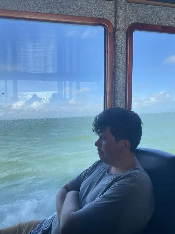 A man sitting by the window inside a ferry, gazing out at the calm sea and clear sky. The reflection on the glass adds depth to the scene, portraying a peaceful and reflective travel experience.