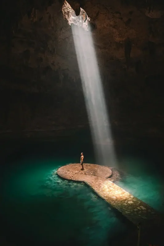 A person standing on a stone platform inside a dark cenote, illuminated by a dramatic beam of sunlight from an opening above. The clear turquoise water surrounding the platform adds to the mystical and awe-inspiring atmosphere of this natural wonder.