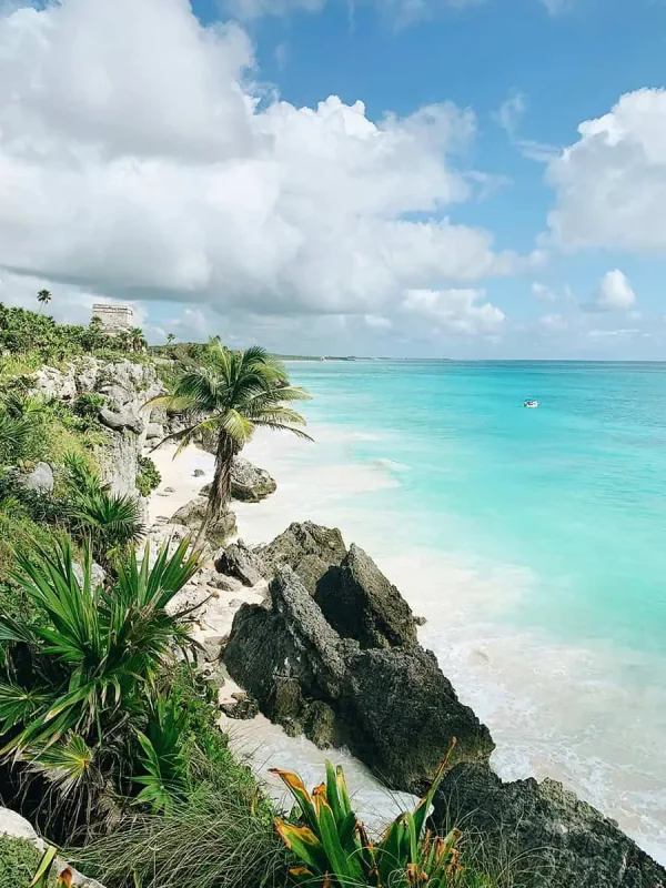 Stunning beach scene in Tulum, Mexico, with clear turquoise waters gently lapping against the white sandy shore. Large rocks and lush greenery add to the picturesque setting, with a historic Mayan structure visible on the cliff, a must-visit on a Mexico and Belize itinerary.