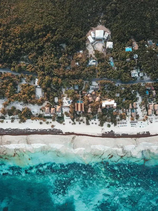 Aerial view of a coastal resort area in tulum with dense greenery, palm trees, and thatched-roof huts lining the pristine sandy beach. The vibrant blue water meets the shore, creating an inviting tropical paradise.