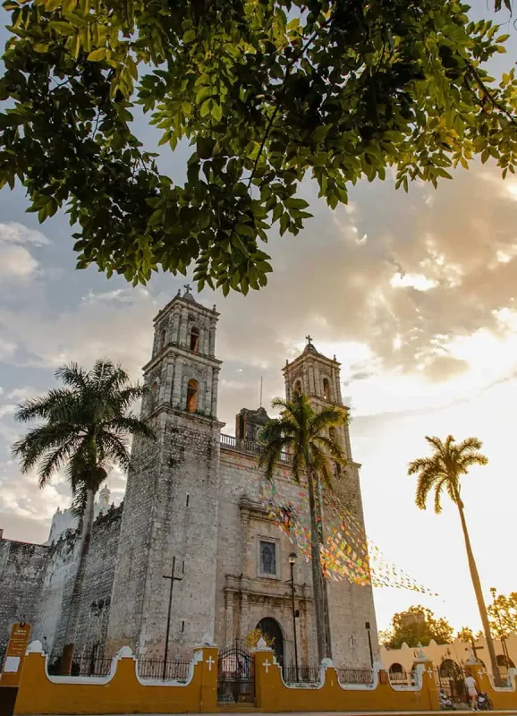 Historic cathedral with twin bell towers framed by palm trees and lush green leaves in Valladolid, Mexico. The late afternoon sun casts a warm glow on the stone facade, capturing the architectural beauty and cultural richness of the region.