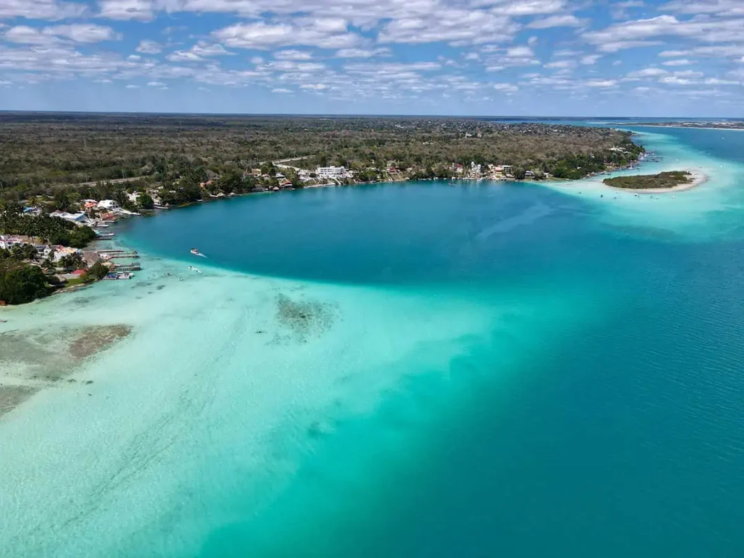 Aerial view of a serene, blue lagoon with a gradient of shallow to deep waters surrounded by lush greenery in Bacalar. The lagoon's calm, clear waters create a peaceful and idyllic setting, perfect for a refreshing swim or relaxation