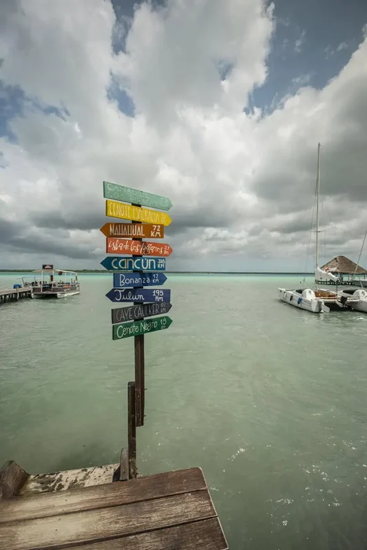 A colorful wooden signpost on a dock by the water, pointing to various destinations such as Cancun, Isla de los Pájaros, and Caye Caulker. The signpost stands against a backdrop of cloudy skies and calm waters, with boats moored nearby, capturing the essence of travel and adventure.