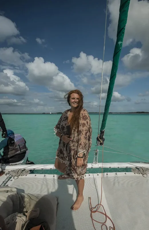 Tasha Amy standing on the deck of a sailboat with turquoise waters and a cloudy sky in the background. She is smiling and holding onto the boat's rigging, enjoying the breeze and the scenic beauty, embodying the joy and excitement of a Mexico and Belize itinerary.