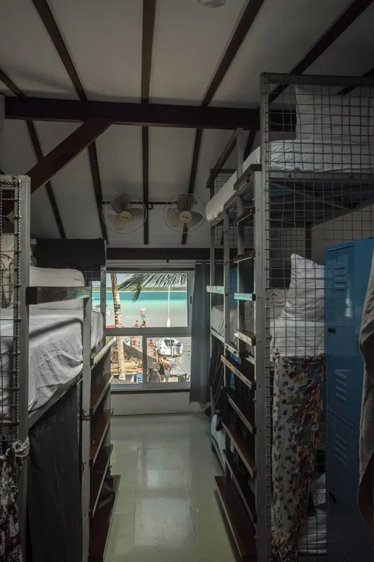 Mexico Belize Guatemala Itinerary: BEST Backpacking Route 28 Inside a yak lakehouse hostel in Bacalar, featuring a row of bunk beds with privacy curtains. The hostel's interior includes high ceilings with fans, and a large window that offers a view of a sunny beach scene outside, providing a unique and inviting atmosphere for travelers.