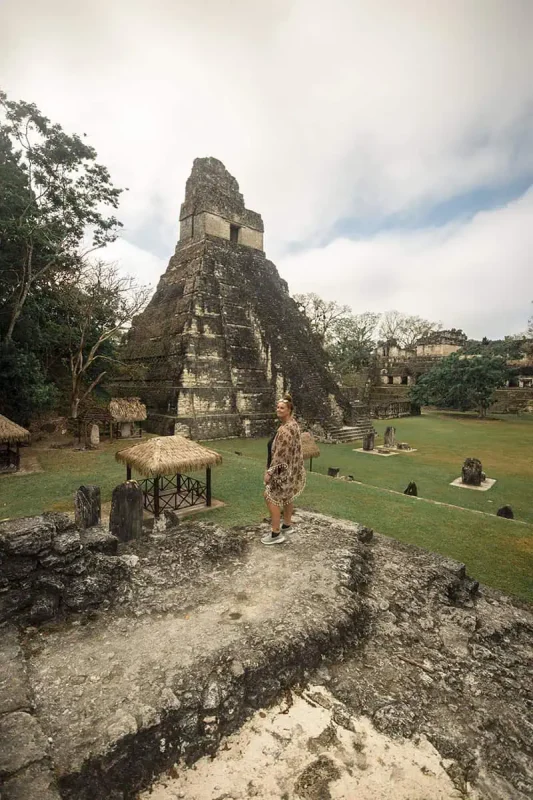Mexico Belize Guatemala Itinerary: BEST Backpacking Route 44 tasha amy standing in front of a massive Mayan temple at Tikal, part of a historical site near Flores. The temple rises dramatically against a backdrop of lush greenery and overcast skies