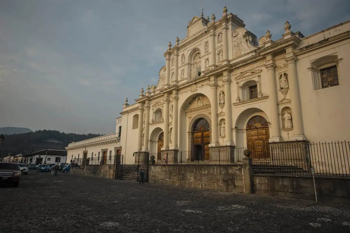 Mexico Belize Guatemala Itinerary: BEST Backpacking Route 61 The historic Cathedral of Antigua, Guatemala, captured in the early evening with a muted sky in the background. The church's elaborate white facade features intricate sculptural details and classical architectural elements. The scene is set on a cobblestone street, enhancing the old-world charm of this colonial city.