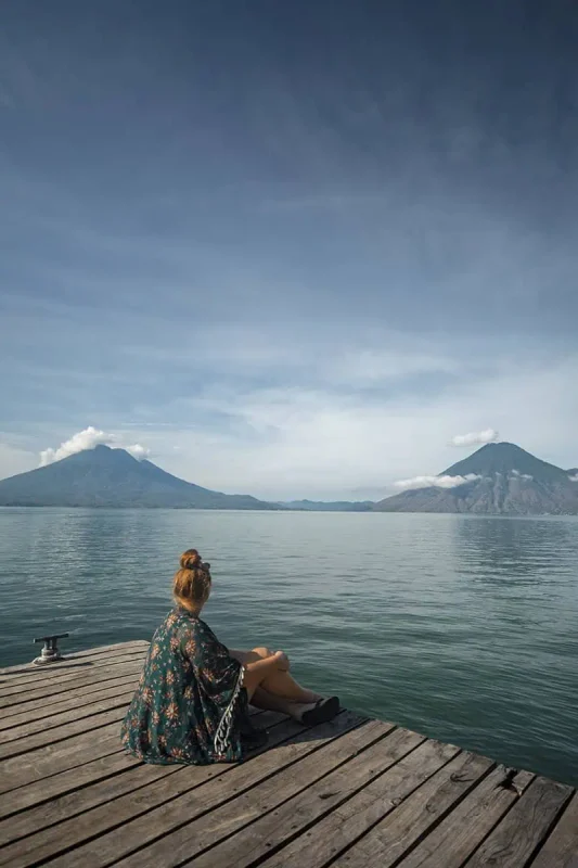 Mexico Belize Guatemala Itinerary: BEST Backpacking Route 66 tasha amy sits on a wooden dock overlooking Lake Atitlán, surrounded by majestic volcanoes under a clear blue sky. Her contemplative pose captures a moment of peace as she gazes out over the calm waters, framed by the natural beauty of Guatemala. This picturesque setting is a serene highlight on the Mexico, Belize, Guatemala itinerary.