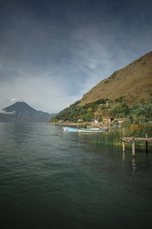 Antigua To Lake Atitlan: By Chicken Bus, Shuttle Or Taxi 14 Lake Atitlán with several boats moored along a pier, set against a backdrop of green hills and a calm lake.