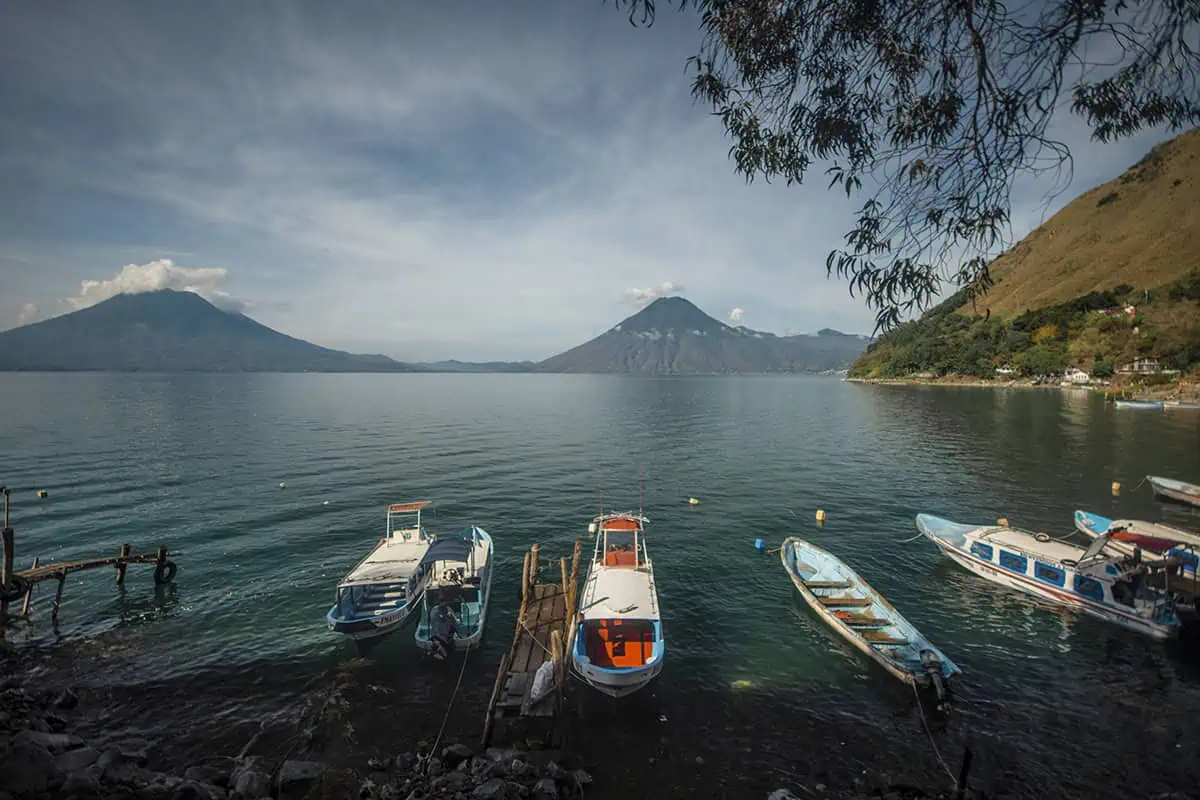 Mexico Belize Guatemala Itinerary: BEST Backpacking Route 68 A panoramic view of Lake Atitlán, captured from a lakeside spot where boats are tied up to a rustic wooden dock. The lake is flanked by towering volcanoes and hills, with trees overhanging the water's edge, presenting a perfect blend of natural elements and serene landscapes.
