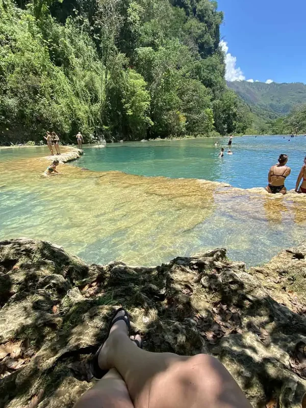 Mexico Belize Guatemala Itinerary: BEST Backpacking Route 52 A first-person perspective showing a woman's legs as she sits on the rocky edge of a natural pool in Semuc Champey. The clear green waters stretch out in front of her, bordered by lush greenery and a vibrant forest scene, reflecting a peaceful and relaxing atmosphere.