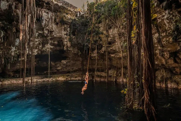 A breathtaking scene of a woman swinging on a rope over a cenote in Tulum, Mexico. This natural sinkhole is surrounded by rugged cliffs adorned with hanging roots and lush vegetation. The water reflects a deep, vivid blue hue, creating a serene yet adventurous atmosphere. this is a must on the mexico belize guatemala itinerary.