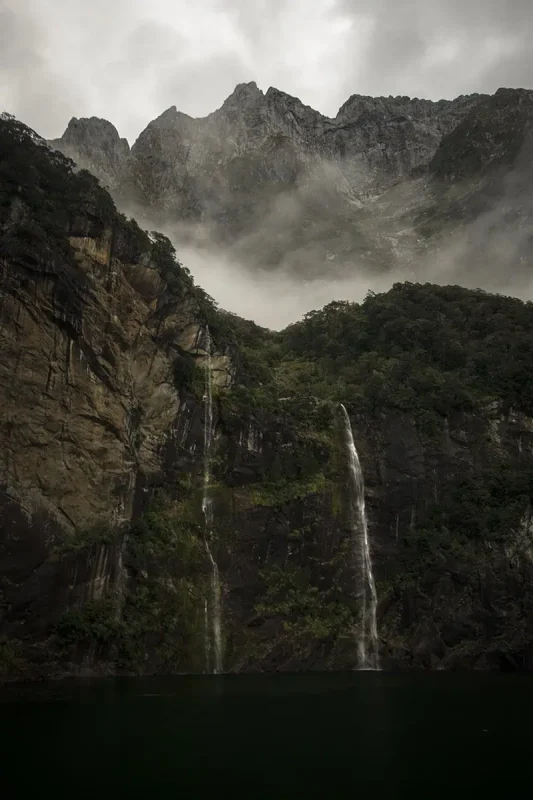 Fiordland & Milford Sound Lord Of The Rings Locations (2025) 5 Twin waterfalls cascade down steep cliffs into dark water with mist clinging to the rocky mountains in Milford Sound, New Zealand