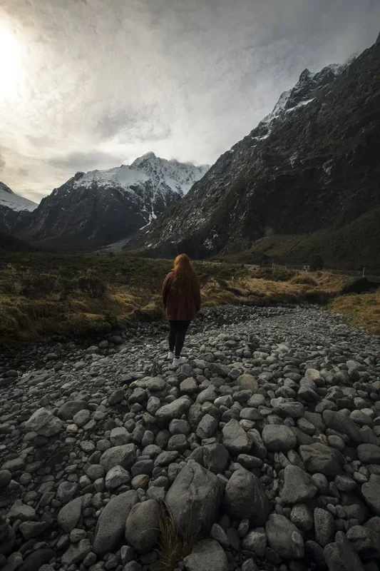 Fiordland & Milford Sound Lord Of The Rings Locations (2025) 6 tasha amy walks across a rocky riverbed surrounded by towering snow-capped mountains in Fiordland, New Zealand