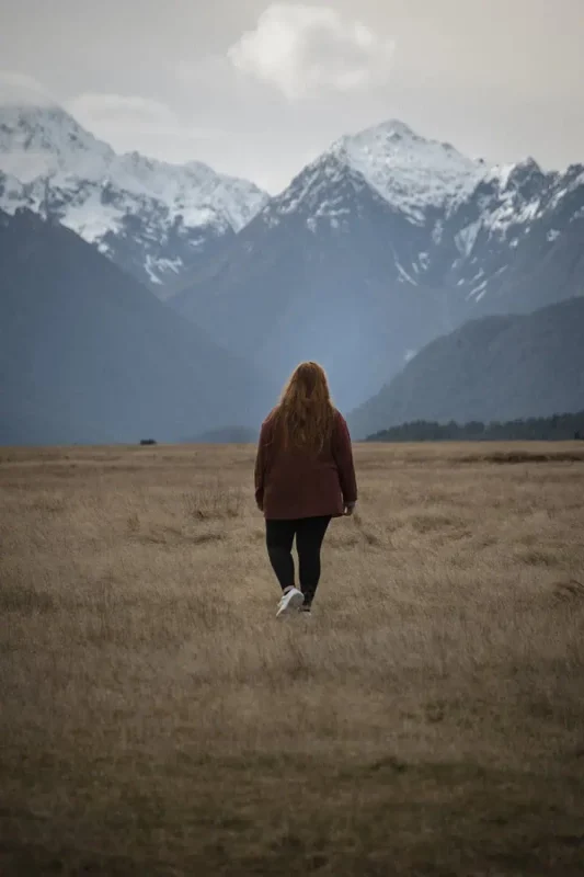 Fiordland & Milford Sound Lord Of The Rings Locations (2025) 20 tasha amy with long hair in a red jacket walks across an open golden field toward sharp snowy peaks in eglinton valley