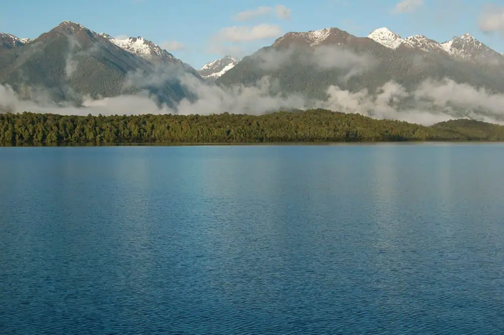 Fiordland & Milford Sound Lord Of The Rings Locations (2025) 9 Calm waters stretch before a line of forest and mountains partly covered in mist at Manapouri Lake