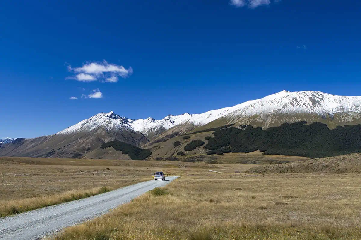 Fiordland & Milford Sound Lord Of The Rings Locations (2025) 7 A car drives along a gravel road toward snow-capped peaks under a blue sky in New Zealand’s Lord of the Rings landscape.