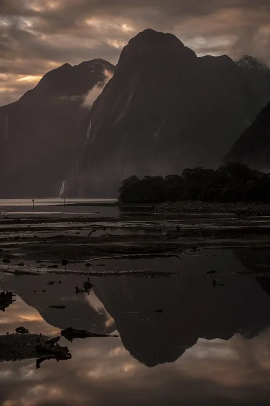 Milford Sound Swing: Where To Find It (2025) 13 Dark shot of cliffs and peaks reflected in the shore, with waterfall streams visible under the sky.