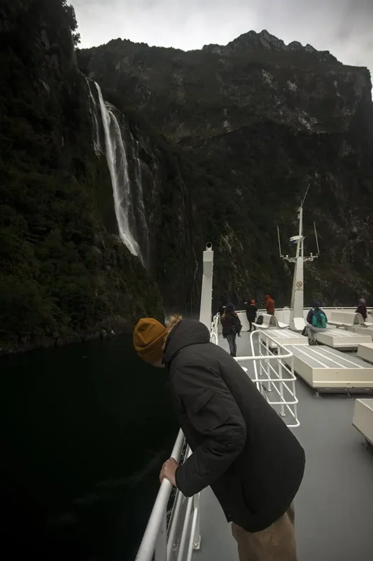 Milford Sound Swing: Where To Find It (2025) 15 A person in a yellow beanie leans over the edge of a boat as it cruises past waterfall cascading down a cliff in Milford Sound.