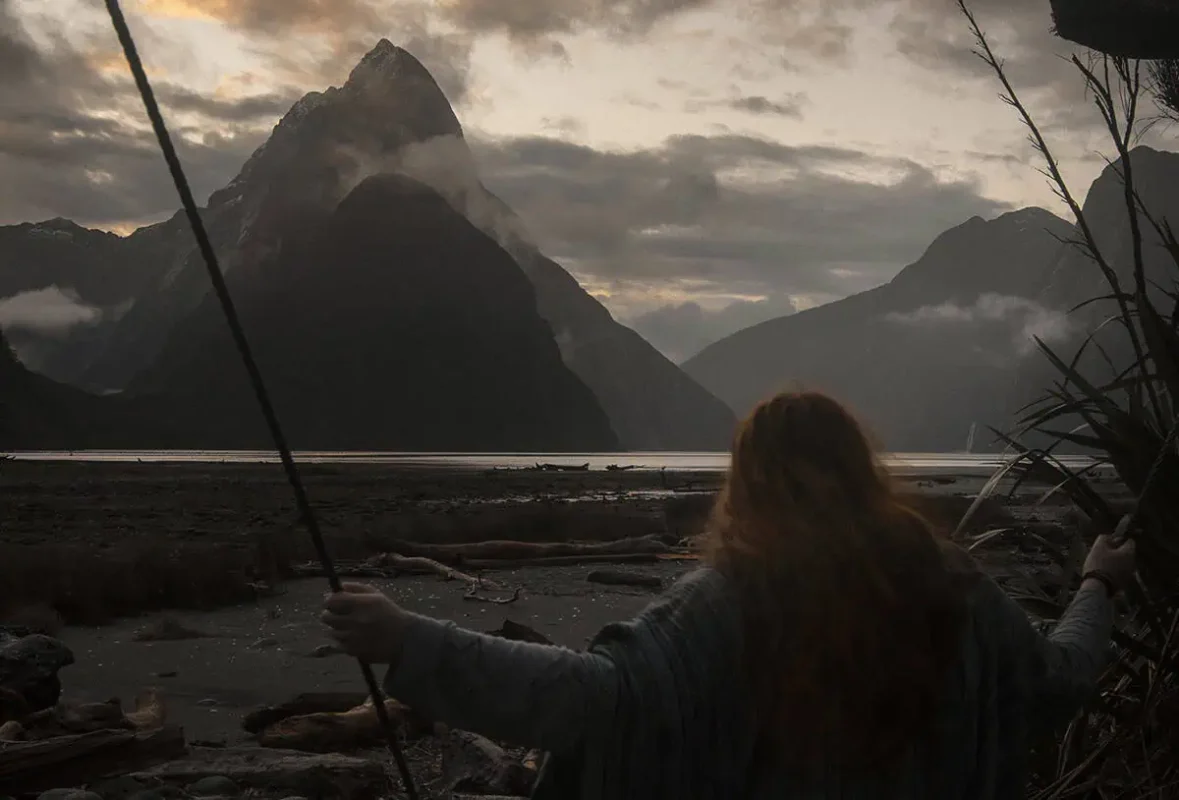 Milford Sound Swing: Where To Find It (2025) 16 tasha amy with long red hair swings at sunset, facing the dramatic silhouette of Mitre Peak in Milford Sound with mist and low clouds rolling over the surrounding mountains.