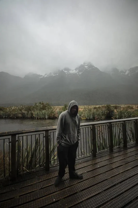 Mirror Lakes: Is It Worth Visiting In Milford Sound, NZ? 7 A person in a gray hoodie stands on a wooden platform overlooking Mirror Lakes in Milford Sound. The platform is surrounded by dense greenery and offers a view of the misty, snow-capped mountains in the background. The gray, cloudy sky and the reflective surface of the lake create a serene and contemplative atmosphere.