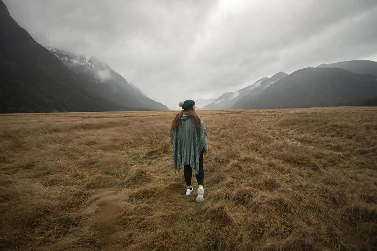 Mirror Lakes: Is It Worth Visiting In Milford Sound, NZ? 15 tasha amy wearing a green poncho and a beanie walks across a vast, golden field towards mist-shrouded mountains in the distance. The wide-open space and towering mountain ranges create a sense of grandeur and adventure, while the overcast sky adds an element of solitude and introspection.