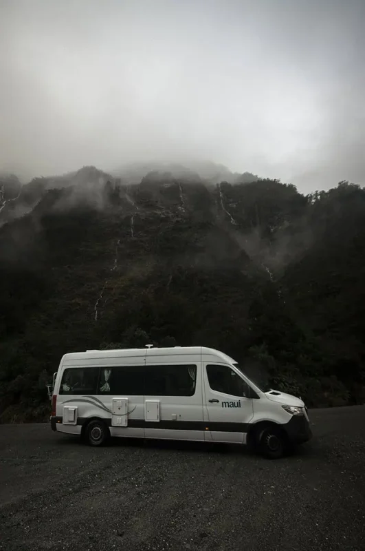 Monkey Creek: BEST Stop On The Way To Milford Sound 12 A white camper van with "Maui" branding is parked on a gravel surface in front of a dark, mist-covered mountainous landscape at Monkey Creek, New Zealand. The rugged mountain is partially obscured by low-hanging clouds and mist, creating an atmospheric, moody scene. The van, with curtains drawn on the windows, stands in stark contrast to the dark, natural surroundings.