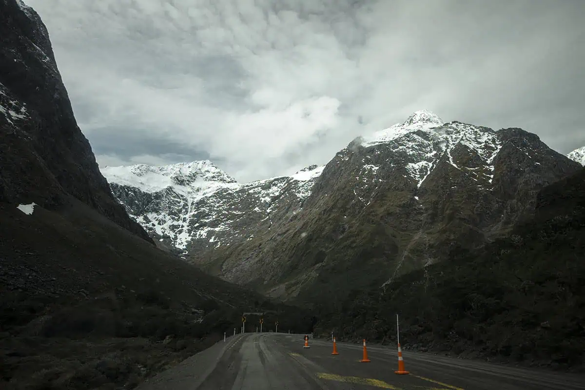 Monkey Creek: BEST Stop On The Way To Milford Sound 8 A scenic road through to Milford Sound in New Zealand, winds through a mountainous landscape with towering, snow-capped peaks. Bright orange traffic cones line the roadside, adding a splash of color to the natural setting. The mountains' rugged surfaces are partially covered in snow, and the sky is filled with thick, low-lying clouds, creating a dramatic backdrop.
