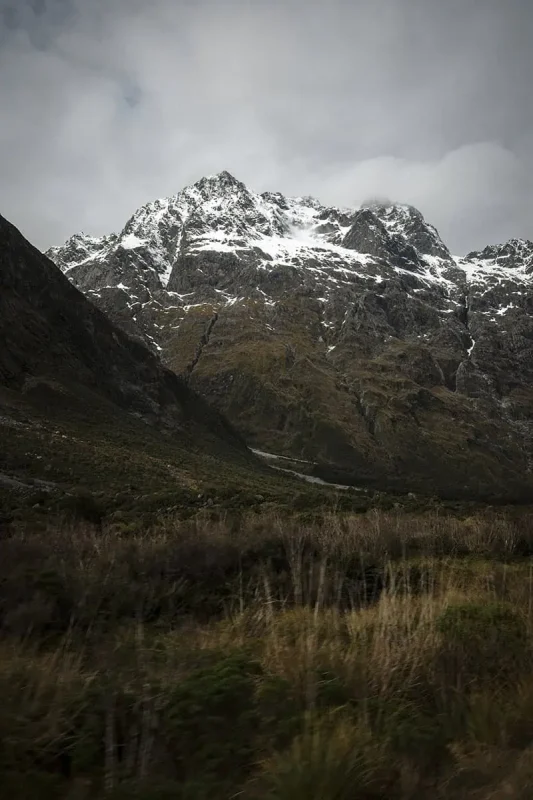 Monkey Creek: BEST Stop On The Way To Milford Sound 20 A striking view of the mountainous terrain at Monkey Creek, New Zealand. The rocky slopes are streaked with patches of snow, contrasting against the green and brown hues of the vegetation below. The cloudy sky overhead adds a moody and atmospheric quality to the scene, emphasizing the raw beauty of the rugged landscape.