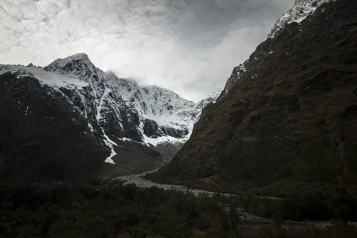 Monkey Creek: BEST Stop On The Way To Milford Sound 15 A panoramic view of Monkey Creek, New Zealand, featuring a wide valley framed by towering, snow-capped peaks. The rocky terrain and lush greenery stretch out towards the mountains, which rise steeply into the cloudy sky. The scene captures the rugged and pristine beauty of this remote location, showcasing its dramatic natural features.